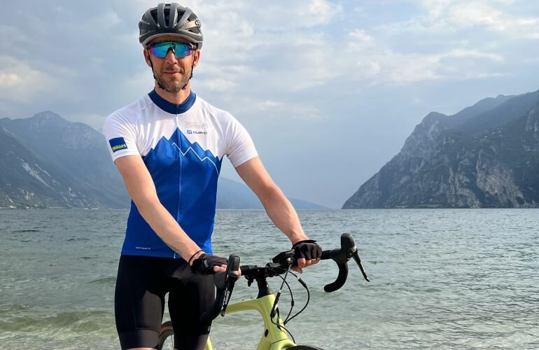 Mann mit Fahrradhelm und Sonnenbrille steht mit Rennrad vor einem See und Bergen im Hintergrund.