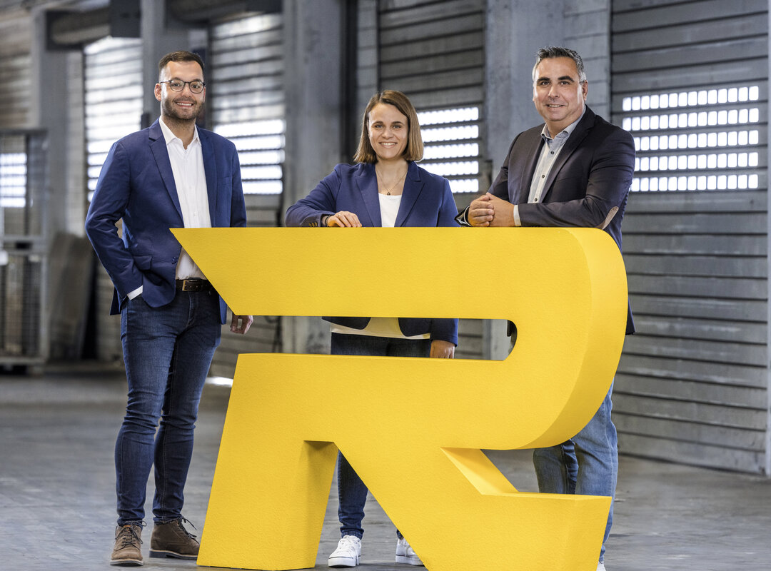 3 Personen stehen hinter dem 'R' von Logo Rieck.