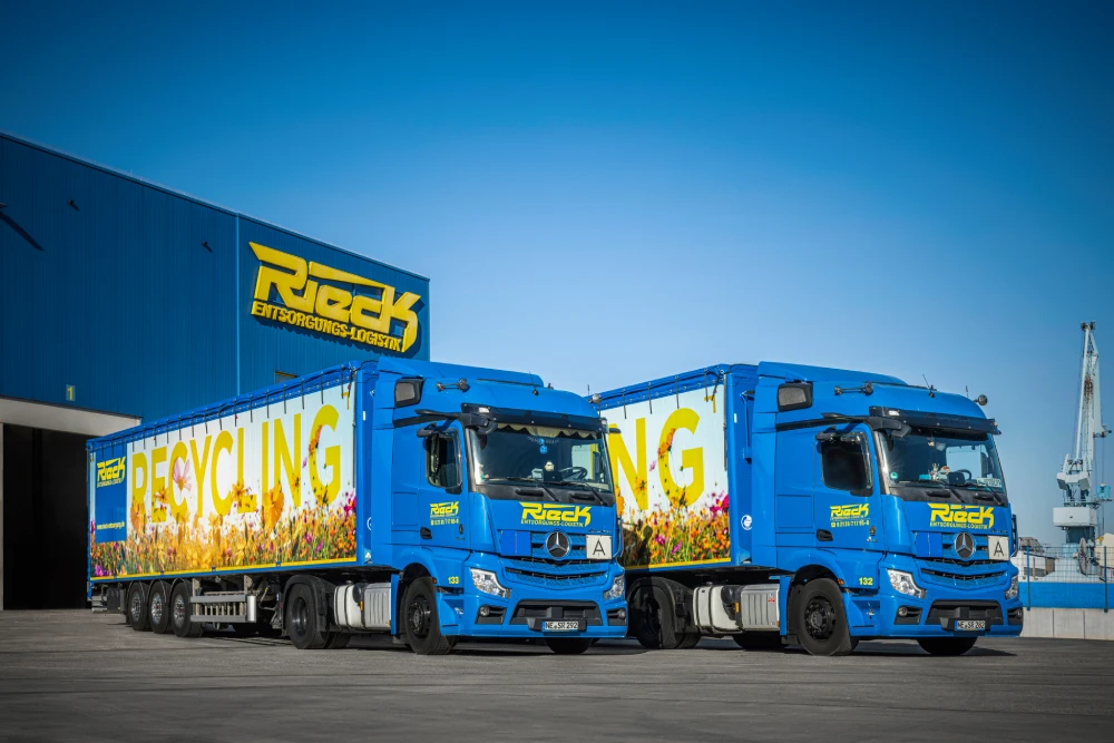 Zwei blaue Recycling-LKWs vor einem blauen Gebäude mit klarem Himmel.