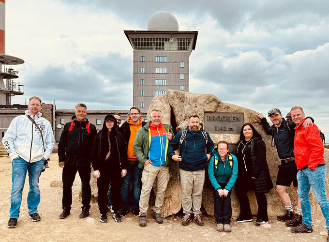 Gruppe von neun Personen vor einem großen Felsen und einem Turm mit Radarkuppel bei bewölktem Himmel.