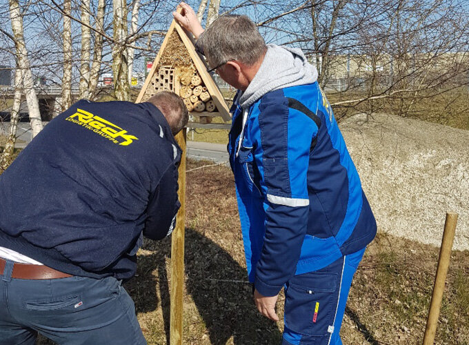 Zwei Männer befestigen ein Insektenhotel an einem Holzpfahl im Freien.