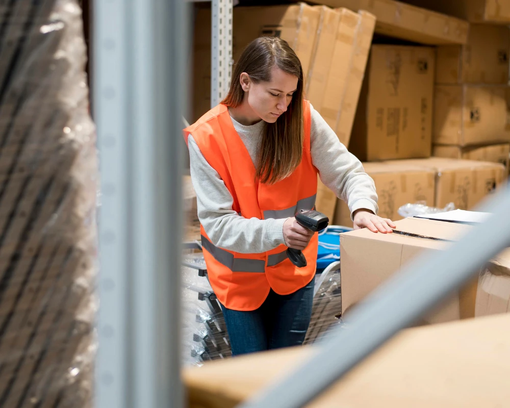 Frau mit oranger Warnweste scannt Barcode auf Karton in Lagerhalle.