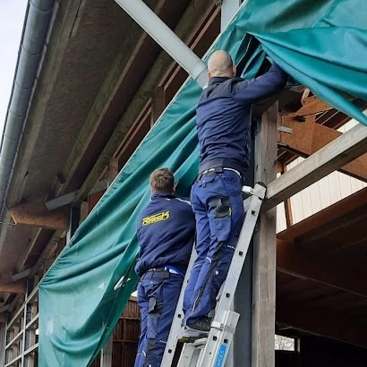Zwei Männer befestigen eine grüne Plane an einem Holzrahmen, einer steht auf einer Leiter.