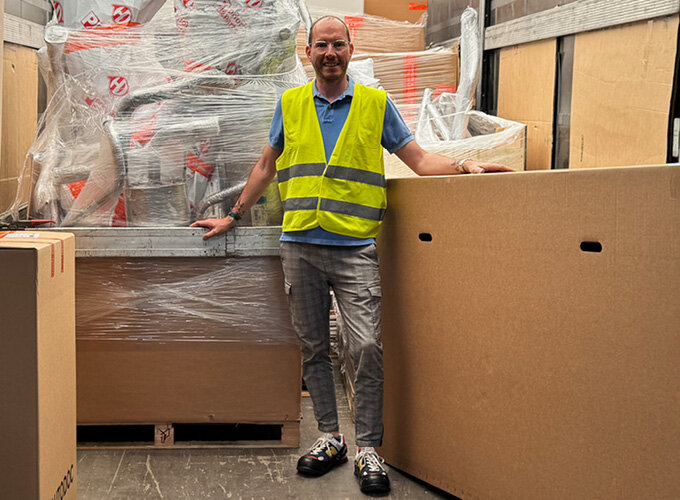 Mann mit gelber Warnweste steht in Lagerhalle zwischen großen Kartons und verpackten Paletten.