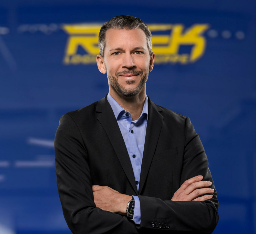 Porträt eines lächelnden Mannes in Anzug vor blauem Hintergrund mit gelbem Schriftzug.