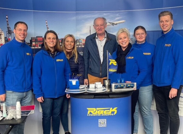 Sechs Personen in blauen Jacken stehen hinter einem Messetisch mit Werbematerialien und einem Logo.